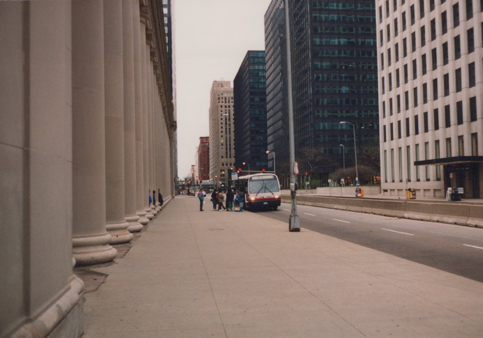 A white and red CTA bus numbered 4566 is stopped on a wide city sidewalk next to large buildings, with a few people standing near it on a cloudy winter day.