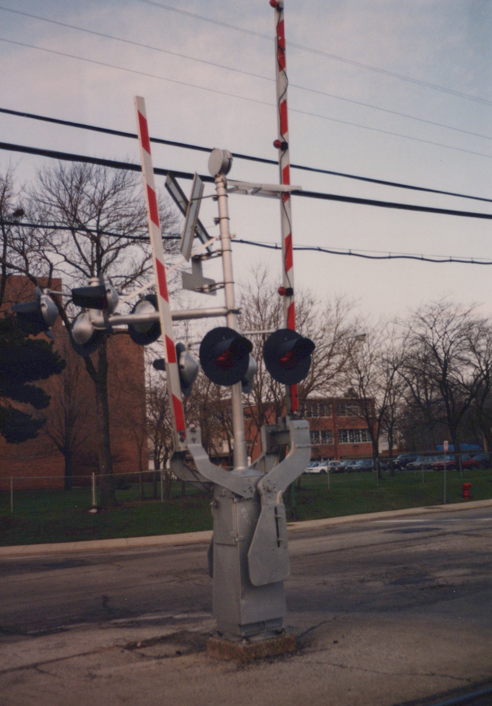 A close-up of a railroad grade crossing signal with red flashing lights and striped gates, located on a street next to bare trees and a brick building in the background.