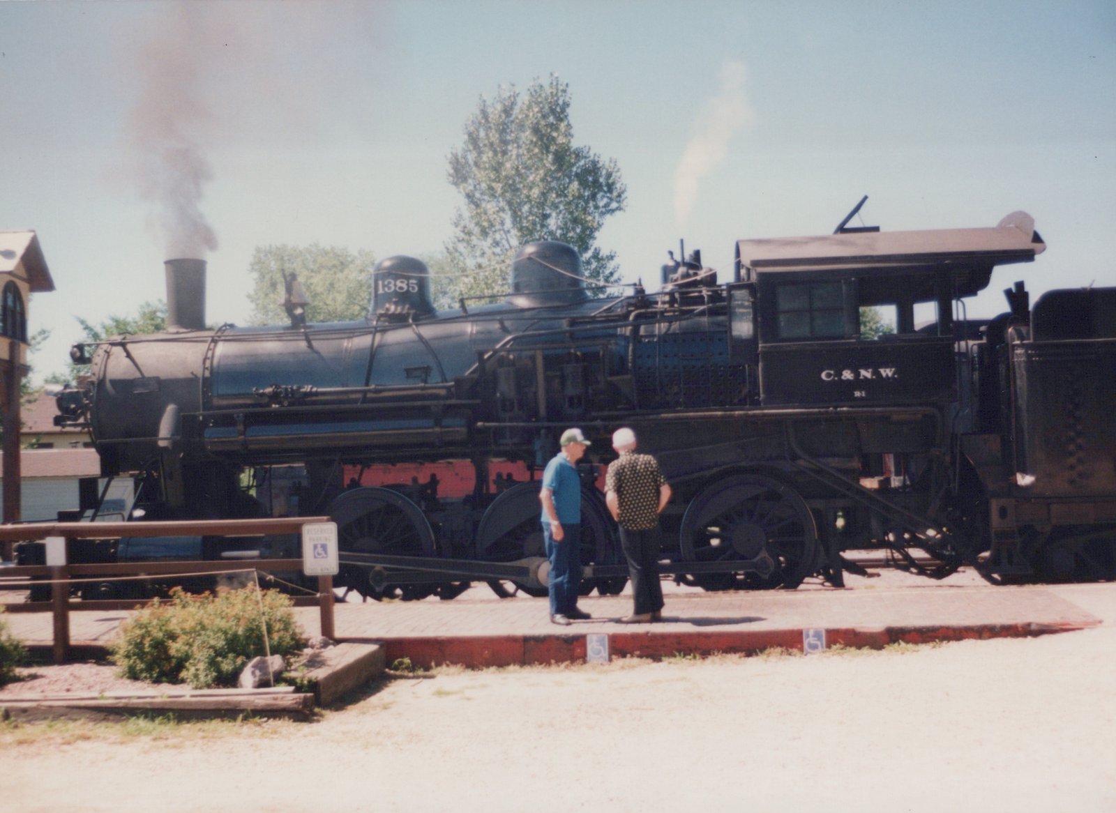 A black steam locomotive numbered 1385 with C.&N.W. lettering stands on tracks next to a platform where two men are observing it, under a clear sky.