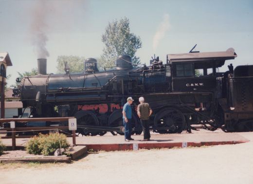 A black steam locomotive numbered 1385 with C.&N.W. lettering stands on tracks next to a platform where two men are observing it, under a clear sky.