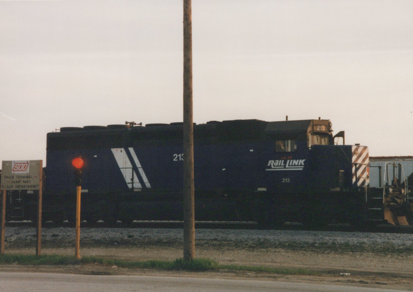 A dark blue I&M Rail Link locomotive numbered 213 is shown from a three-quarter front view in a railroad yard, with a red signal light and a sign for a truck terminal and hump yard visible nearby.