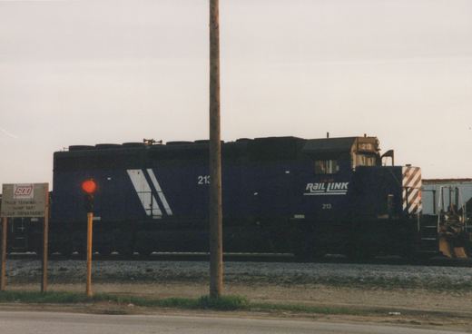 A dark blue I&M Rail Link locomotive numbered 213 is shown from a three-quarter front view in a railroad yard, with a red signal light and a sign for a truck terminal and hump yard visible nearby.