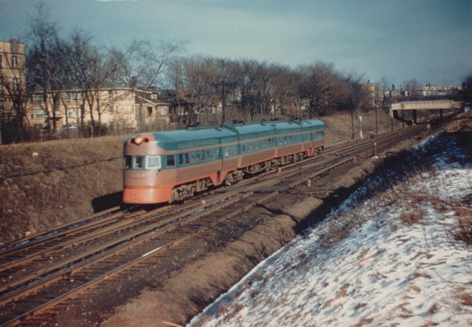 A single-unit, streamlined railcar with a teal, orange, and silver paint scheme is shown on railroad tracks next to a snow-covered embankment and bare trees in the winter.