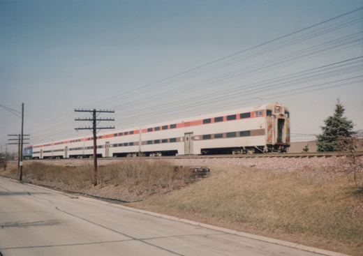 A side view of a Metra train with bi-level Superliner passenger cars moving along tracks beside a road, with utility poles and a clear sky in the background.