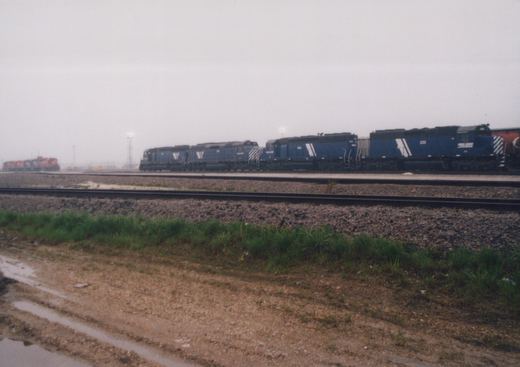 A side view of a blue Wisconsin Central Ltd. locomotive numbered 222 leading a line of locomotives on multiple tracks, with a muddy foreground and an overcast sky.
