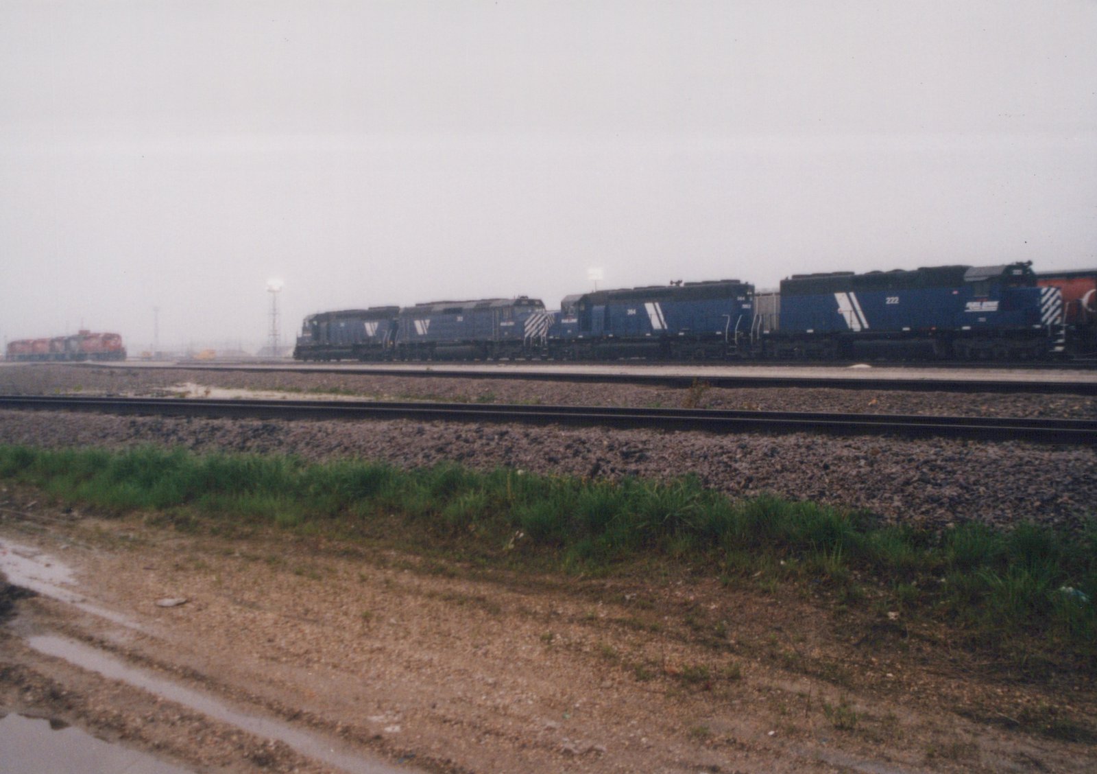 A side view of a blue Wisconsin Central Ltd. locomotive numbered 222 leading a line of locomotives on multiple tracks, with a muddy foreground and an overcast sky.