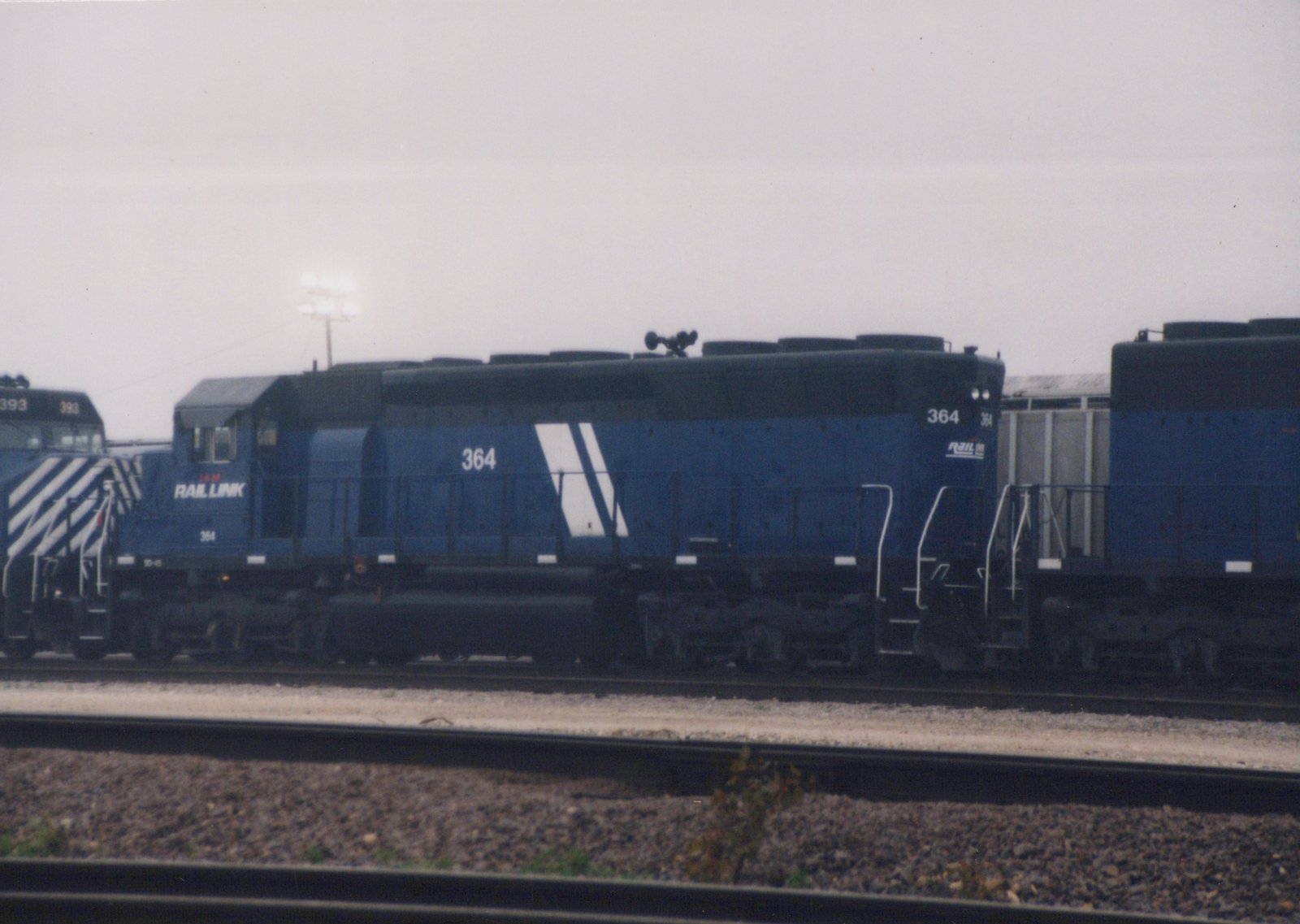 Side view of a blue Rail Link diesel locomotive numbered 364 on a track in a rail yard, with another locomotive marked 393 in the background under an overcast sky.