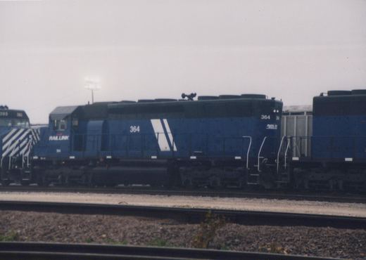 Side view of a blue Rail Link diesel locomotive numbered 364 on a track in a rail yard, with another locomotive marked 393 in the background under an overcast sky.