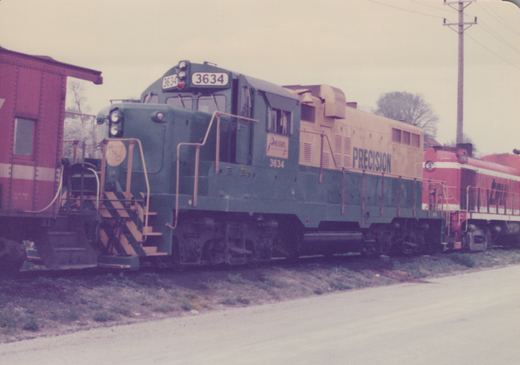 A green and tan diesel locomotive with "PRECISION" and number 3634 on its side, parked on a railway track next to other railcars.