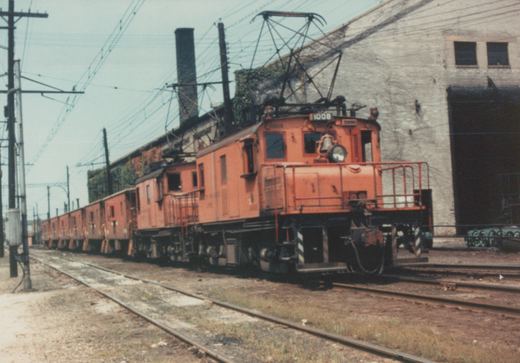 An orange electric locomotive numbered 11008 with a pantograph, pulling a long line of matching orange boxcars in a railroad yard under overhead wires.