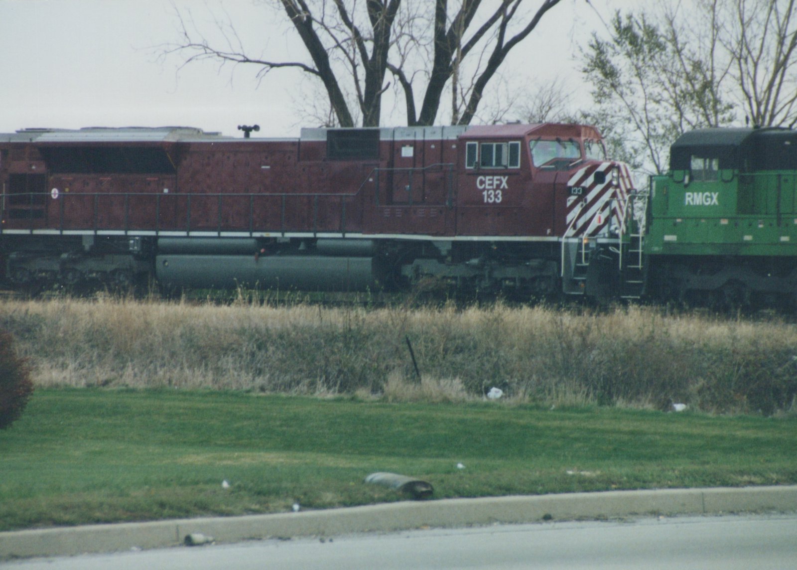 A red CEFX 133 diesel locomotive and a green RMGX unit are on parallel tracks with bare trees and grass in the background.