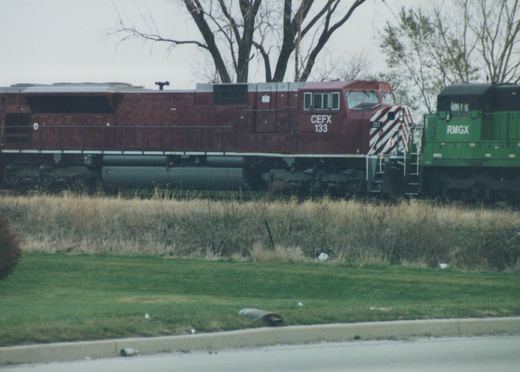 A red CEFX 133 diesel locomotive and a green RMGX unit are on parallel tracks with bare trees and grass in the background.