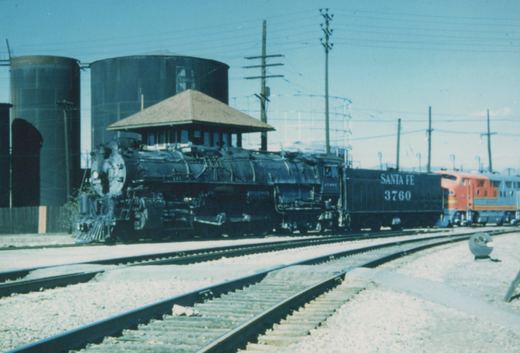 A black Santa Fe steam locomotive numbered 3760 is on the tracks in a yard with a red and silver diesel locomotive visible behind it.