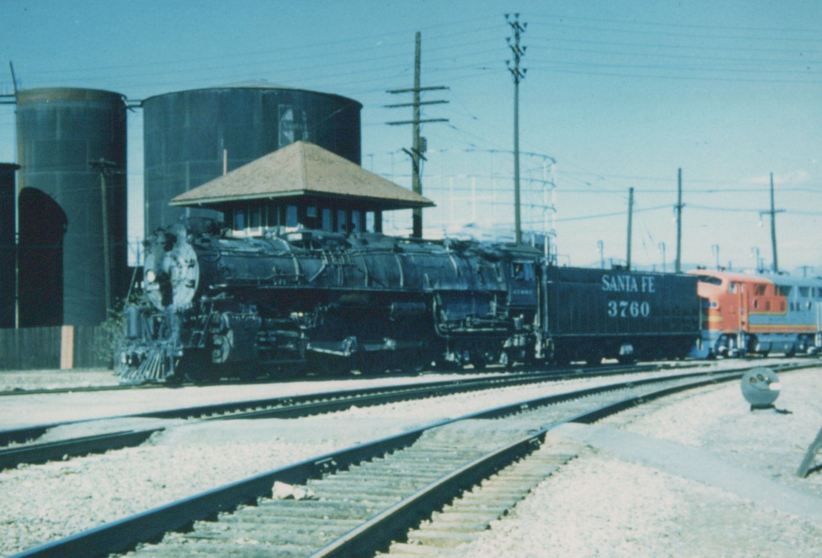 A black Santa Fe steam locomotive numbered 3760 is on the tracks in a yard with a red and silver diesel locomotive visible behind it.