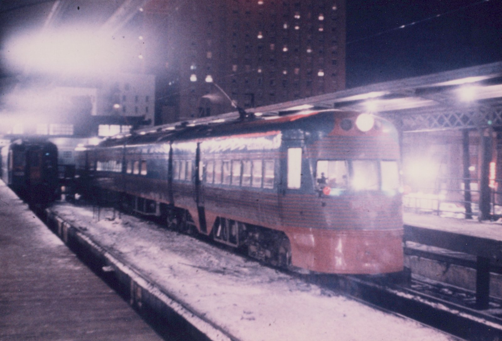 A vintage electric interurban car with a streamlined body and trolley pole is parked at a snowy station platform on an elevated railway at night in a city setting.