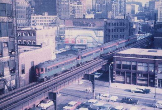 An elevated view of a green and red electric train on a steel viaduct in a city, with 1950s cars on the street below.