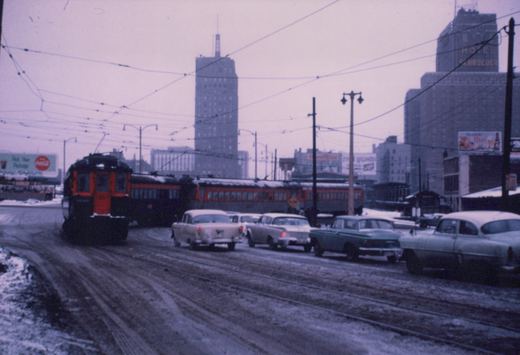 A red and black interurban train on a snowy city street, with vintage cars and tall buildings in the background.
