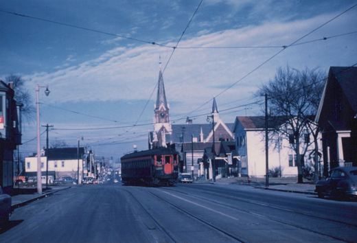 A vintage dark green and red interurban car runs on streetcar tracks down a tree-lined street with a church steeple in the background on a cloudy winter day.