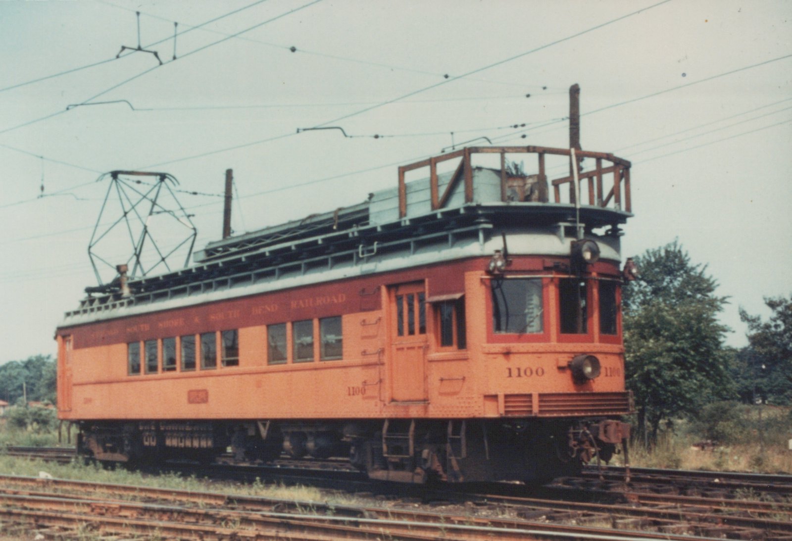 A vintage orange and maroon interurban railcar, number 1100 of the Chicago South Shore and South Bend Railroad, sits on tracks with overhead wires and trees in the background.