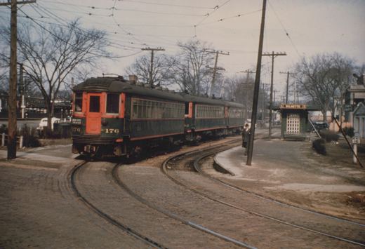 A dark green and orange interurban train with the number 176 is stopped at a small station platform in a bare-treed winter scene, with a man standing nearby.