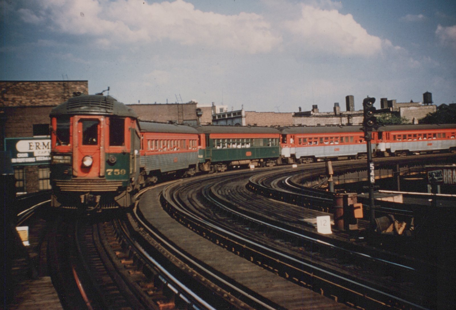 A red and green Chicago North Shore and Milwaukee interurban train, led by car 759, on elevated tracks with city buildings in the background under a partly cloudy sky.