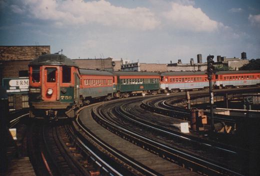 A red and green Chicago North Shore and Milwaukee interurban train, led by car 759, on elevated tracks with city buildings in the background under a partly cloudy sky.