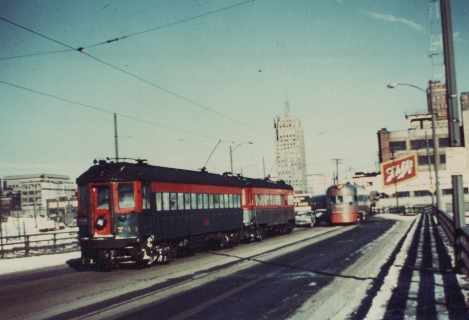 A red and green streamlined interurban train car with the number 741 on the front, moving along a snow-lined city street with overhead wires and a 'Schlitz' billboard in the background.