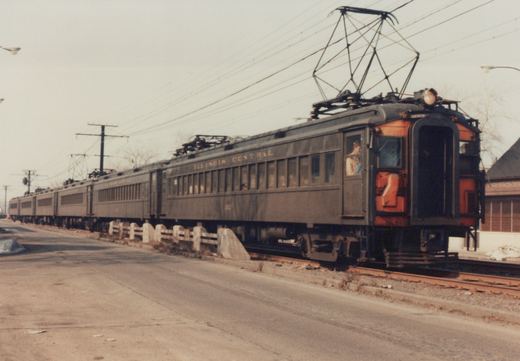 A vintage dark green and orange electric train with the words ILLINOIS CENTRAL on its side, running on tracks next to a street in a winter scene.