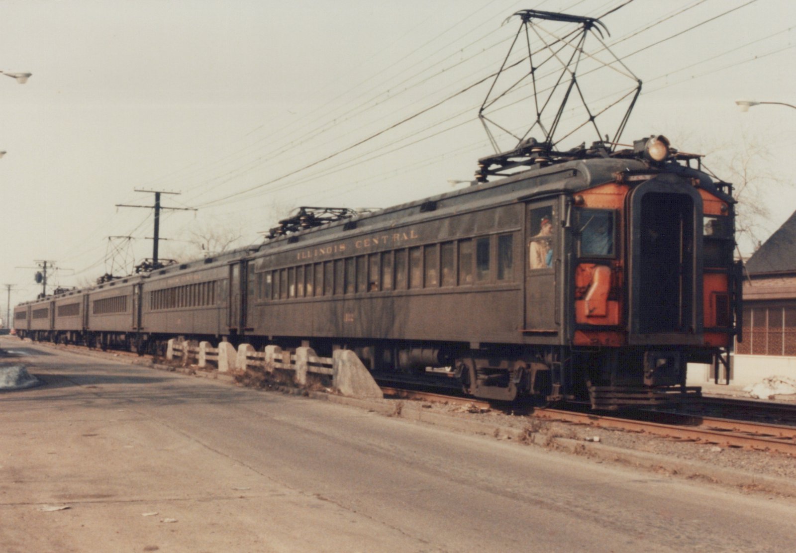 A vintage dark green and orange electric train with the words ILLINOIS CENTRAL on its side, running on tracks next to a street in a winter scene.
