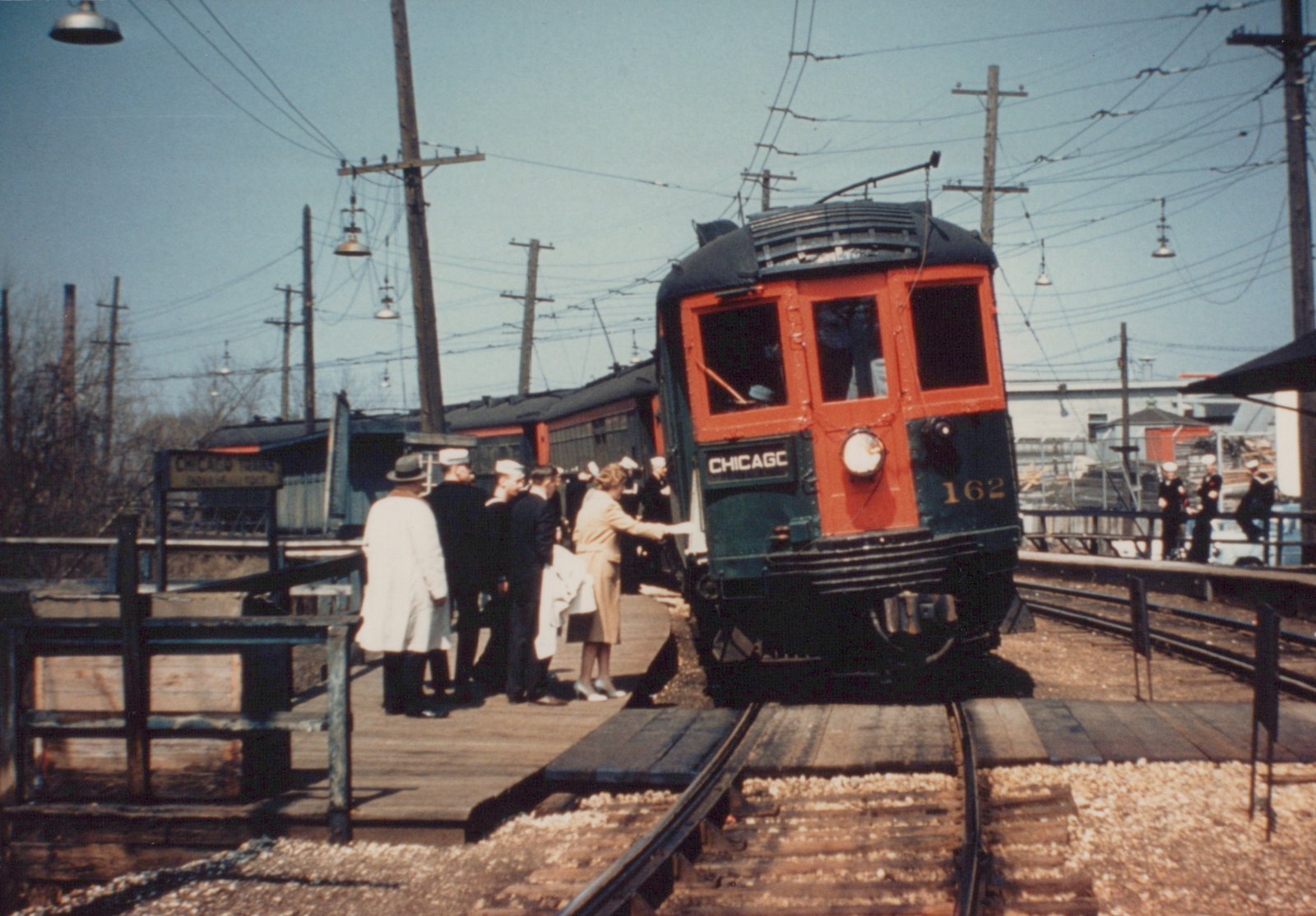 A vintage photograph of a green and orange interurban railcar, number 162, stopped at a wooden station platform with people boarding, under a clear sky with overhead wires.