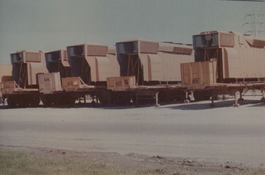 A rail yard scene with several flatcars carrying large beige equipment boxes, numbered 653, 655, 656, and 657, under a bright sky with a power line in the background.