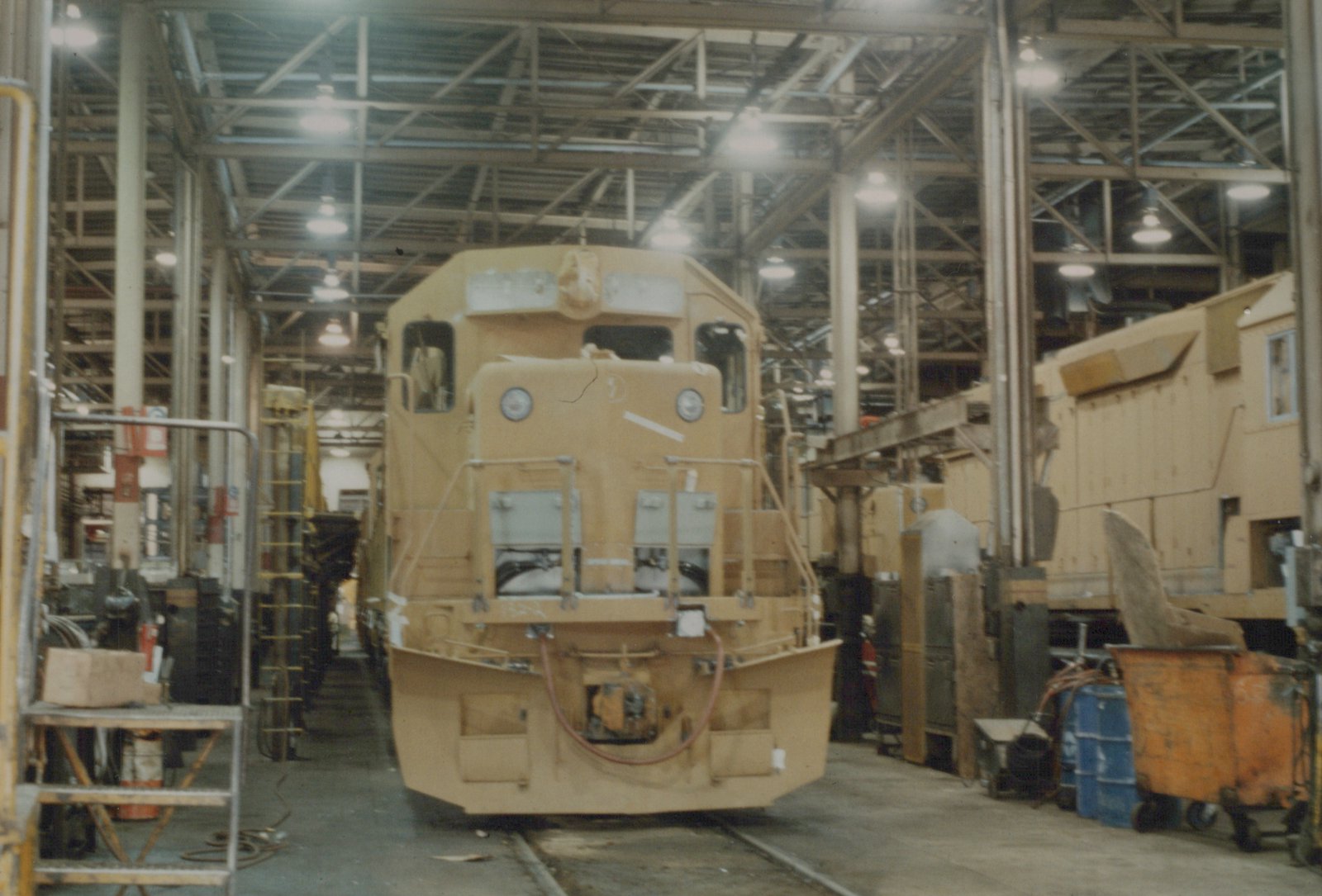 Front view of a yellow SW1200 diesel locomotive inside a large industrial maintenance shop with exposed steel beams and overhead lighting.