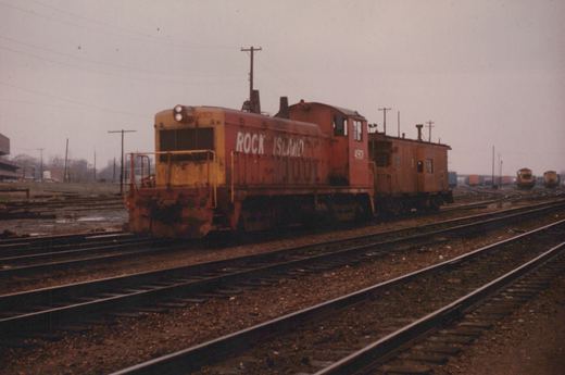 A red and yellow Rock Island freight locomotive numbered 4901 is parked on a set of tracks next to a matching caboose, with other trains visible in the background under a gray sky.