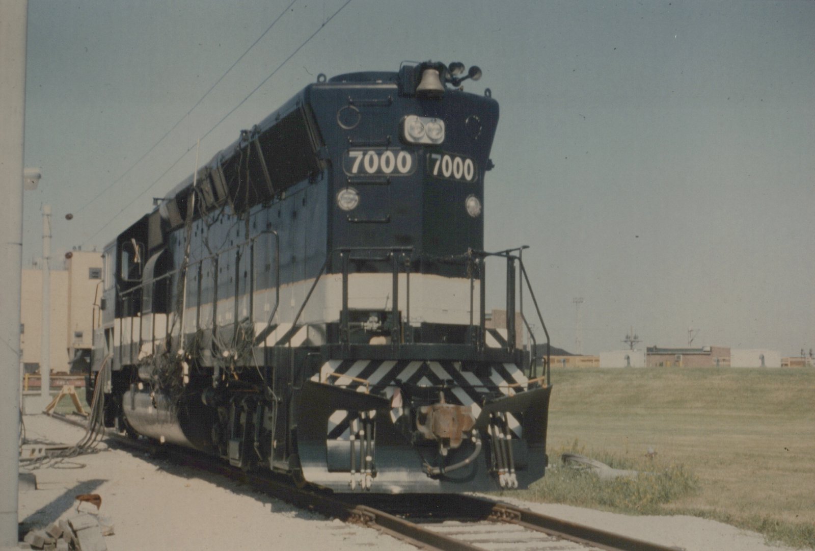 A dark blue and white diesel locomotive, number 7000, is shown at a three-quarter front angle, parked on tracks in a railroad yard with grass and buildings in the background.