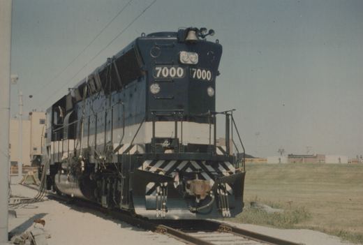 A dark blue and white diesel locomotive, number 7000, is shown at a three-quarter front angle, parked on tracks in a railroad yard with grass and buildings in the background.