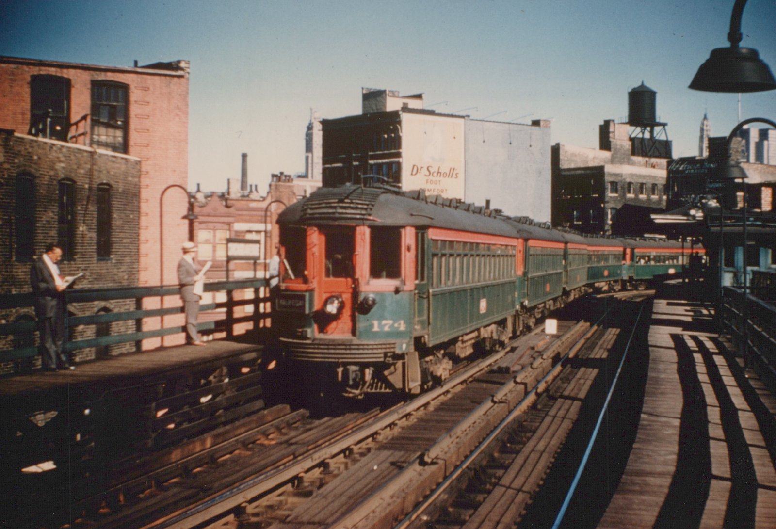 A red and green interurban train, identified as car 174, sits at a wooden station platform under a clear blue sky, with brick buildings and a Dr. Scholl's billboard in the background.