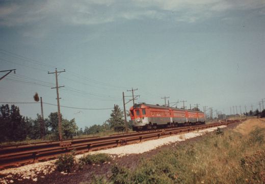 An orange and silver electric train with a pantograph on its roof travels on a two-track line surrounded by grass and utility poles under an overcast sky.