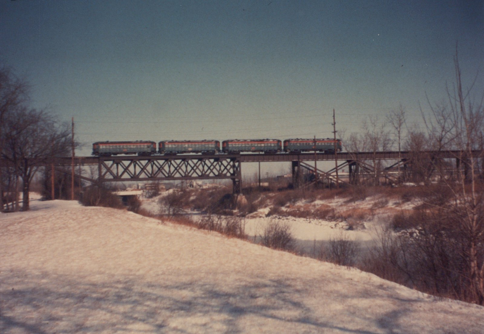 A train crosses a metal trestle bridge over a snowy, brushy valley on a cloudy winter day.