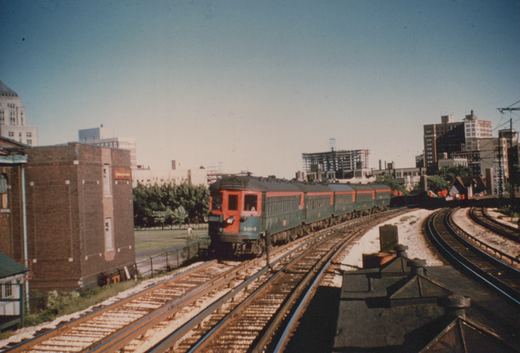 A red and green electric train with the number 104 on the front travels on tracks through a city, with buildings and trees in the background under a clear blue sky.