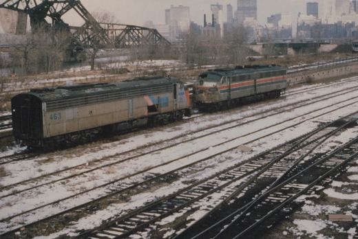 Two Amtrak diesel locomotives on parallel railroad tracks in a snow-covered yard, with a city skyline and a large bridge in the background.