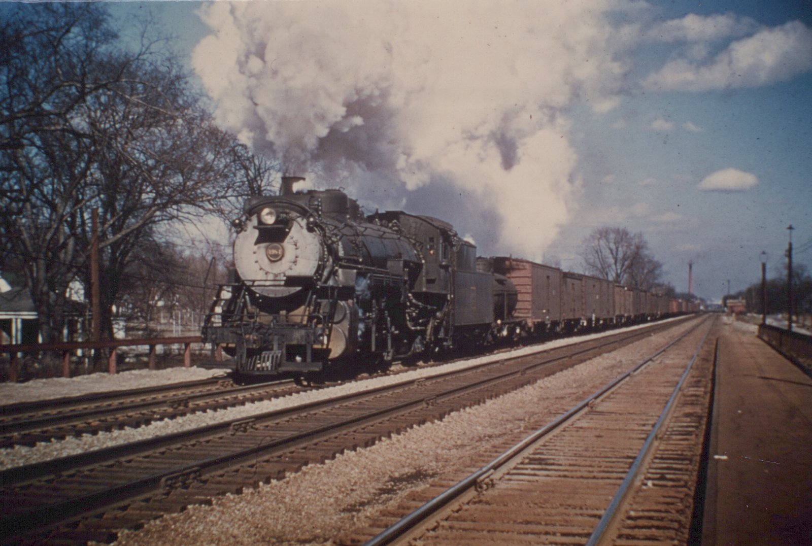 A black steam locomotive with the number 1984 on its front, emitting white smoke, is shown pulling a long line of freight cars along multiple tracks next to a station platform under a blue sky with bare trees nearby.
