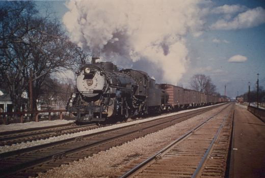 A black steam locomotive with the number 1984 on its front, emitting white smoke, is shown pulling a long line of freight cars along multiple tracks next to a station platform under a blue sky with bare trees nearby.