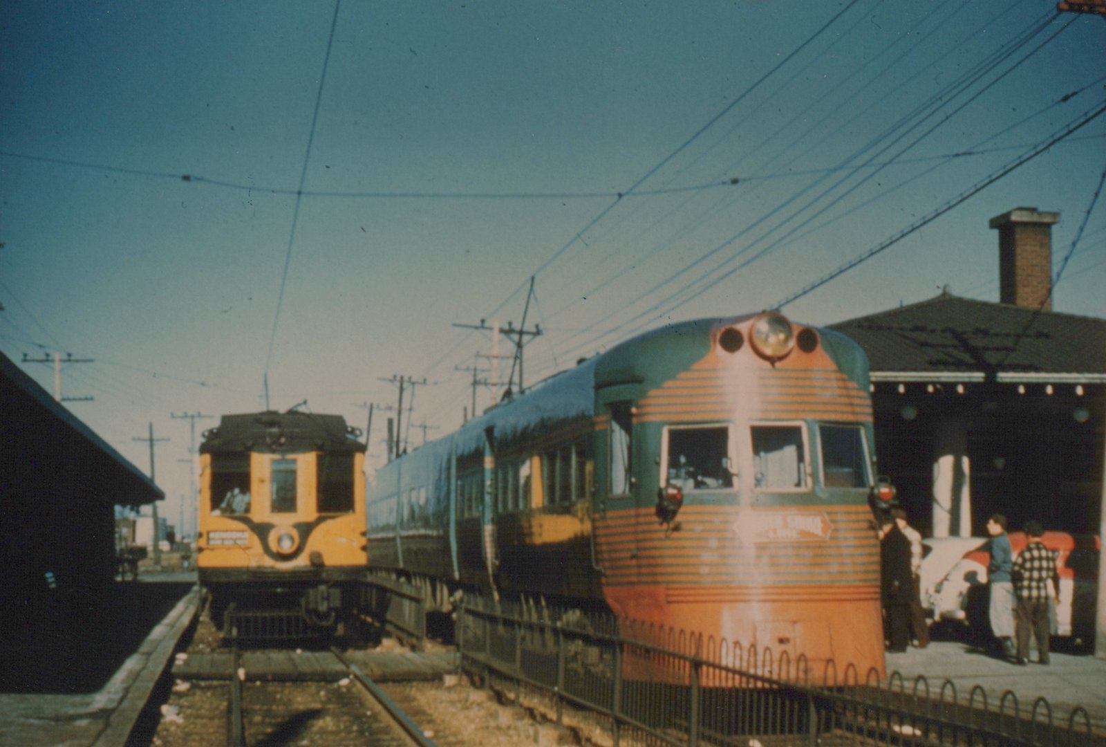 A green and orange streamlined electric train at a station with people on the platform and overhead wires above.