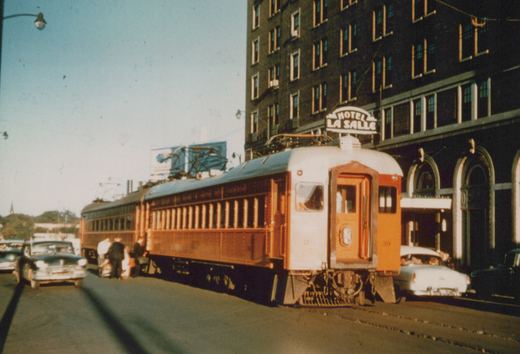 A vintage orange and white interurban train car numbered 39 is stopped on a city street next to a large brick building with a sign reading 'HOTEL LA SALLE'. Several people are boarding, and classic automobiles are parked nearby under a clear blue sky.