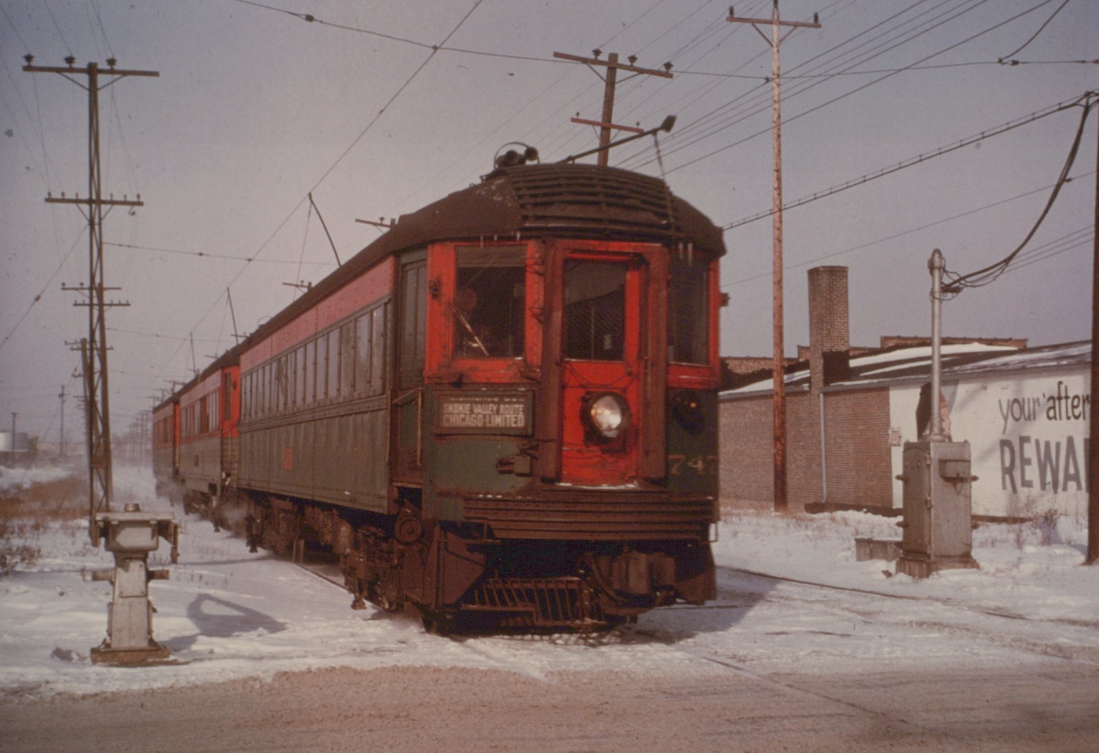 A red and green interurban car with the number 747 and a sign reading 'Skokie Valley Route Chicago-Limited' is stopped on a snow-covered track with overhead wires and utility poles in the background.