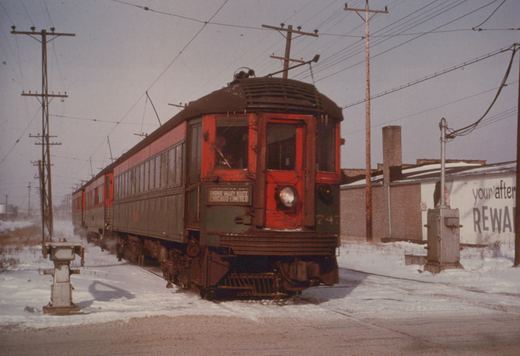 A red and green interurban car with the number 747 and a sign reading 'Skokie Valley Route Chicago-Limited' is stopped on a snow-covered track with overhead wires and utility poles in the background.