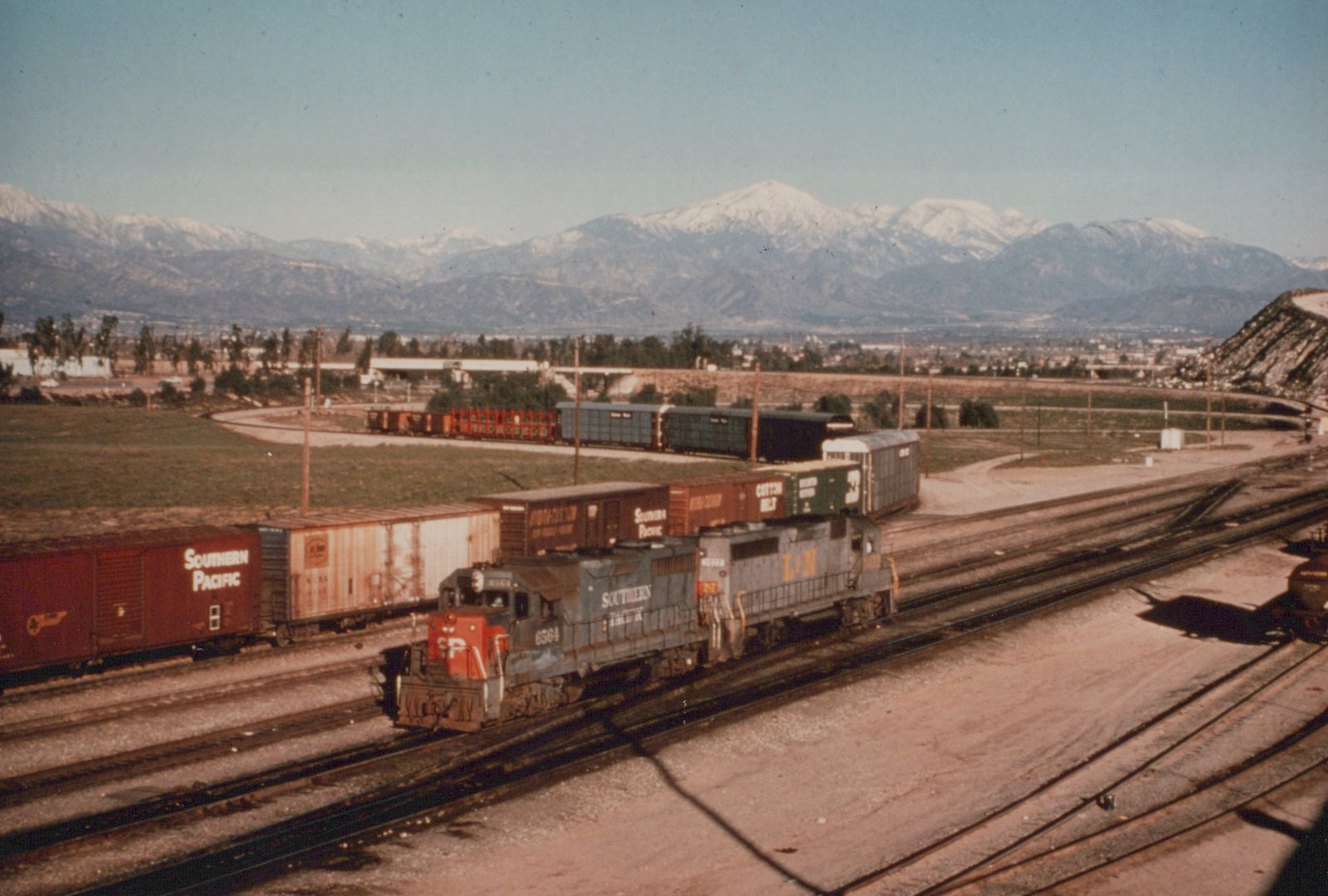 Two locomotives, one Southern Pacific and one Louisville and Nashville, pull a line of freight cars through a yard with mountains in the distance.