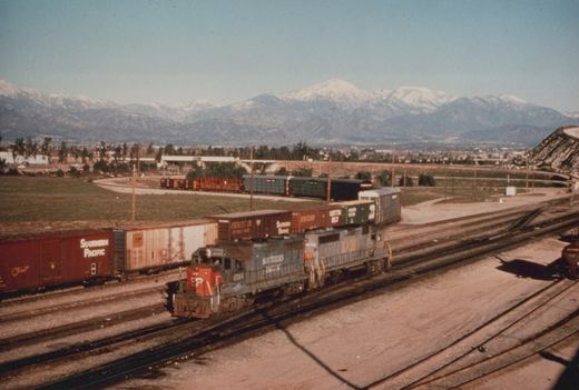Two locomotives, one Southern Pacific and one Louisville and Nashville, pull a line of freight cars through a yard with mountains in the distance.