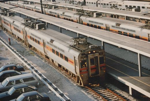 A silver, orange, and red electric train with pantographs, labeled 'Chicago South Shore and South Bend Railroad', is at an elevated station platform. Parked cars are visible alongside the tracks.