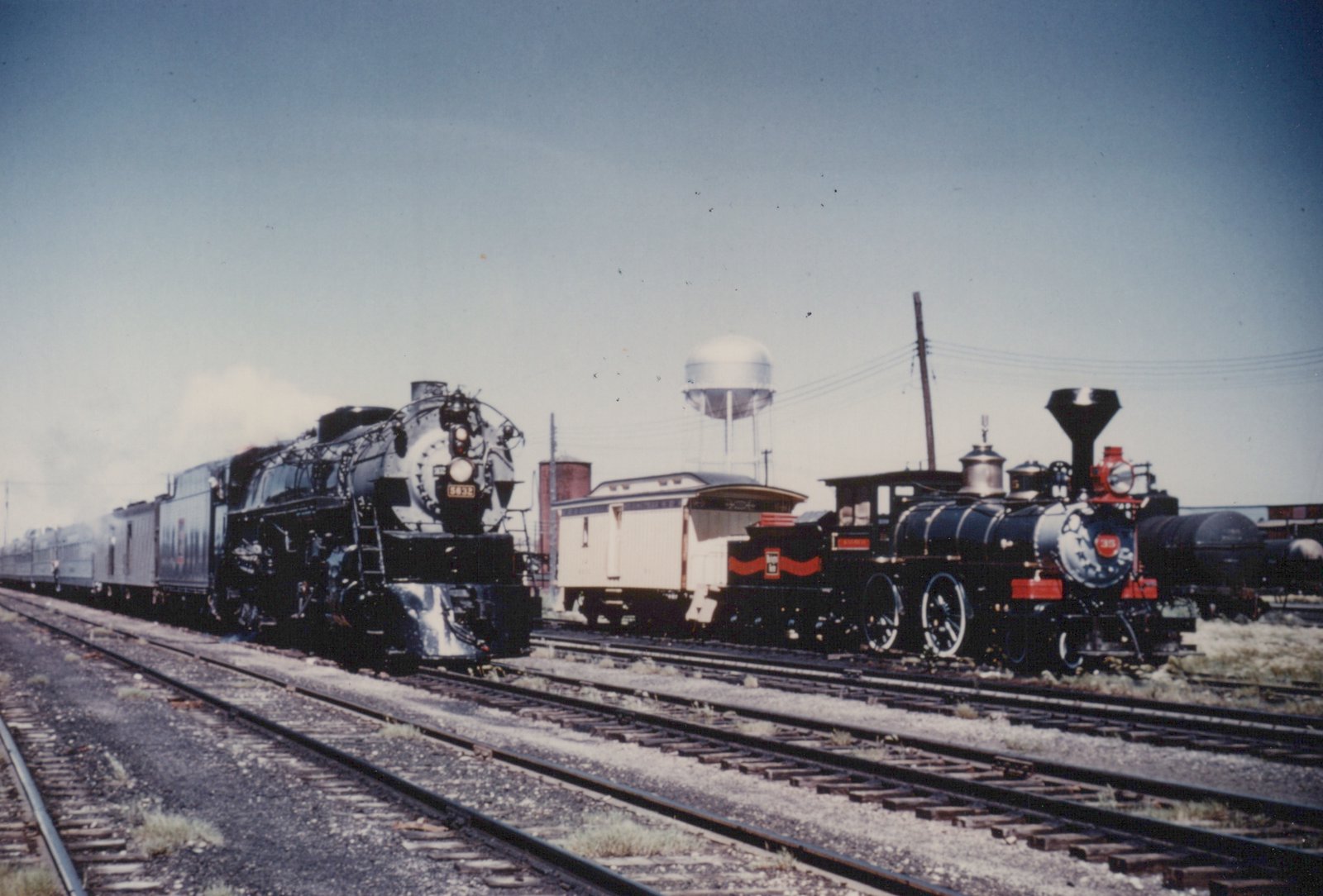 Two steam locomotives, one large and black with the number 6032 and one small and black with the number 35, are on parallel tracks in a railroad yard with a white passenger car and a water tower in the background.